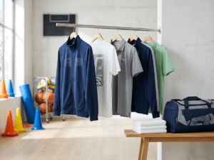 Hochwertige Vereinsausstattung mit bedruckten Trainingsjacken, T-Shirts und Polos auf einer Kleiderstange sowie einer Sporttasche in einer modernen Sportumgebung